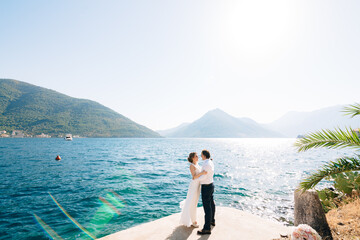 Fototapeta na wymiar The bride and groom hug on the pier in the Bay of Kotor against the background of mountains and the shining sea