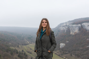 A young dark-haired woman on the background of an autumn landscape. Cloudy gray foggy view from the mountain. A happy smiling girl in a green coat. Natural background, concept of hiking, ecotourism