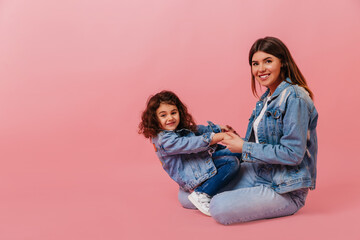 Obraz na płótnie Canvas Glad young woman sitting with daughter on pink background. Studio shot of charming mom and little girl in denim jackets.