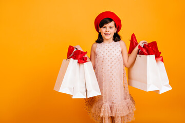 Joyful girl in elegant dress smiling on yellow background. Cute child holding store bags and looking at camera.