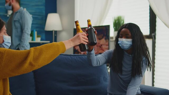 Multiracial group of friends with masks enjoying a beer during new normal party in living room respecting social distance to prevent virus spread. Diverse people enjoying free time in global pandemic