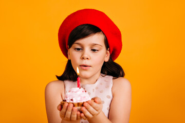 Studio shot of brunette kid making birthday wish. Charming child in beret holding cake with candle.