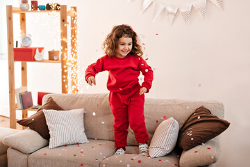 Carefree kid in pajama jumping on sofa. Brunette little girl standing on couch.