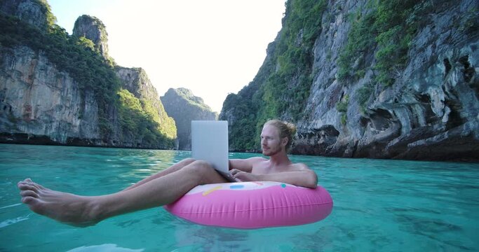 Young freelancer caucasian male floating on inflatable donut in the sea and remote working anywhere on his laptop computer connected to office and relax by seeing beautiful view on summer vacation.
