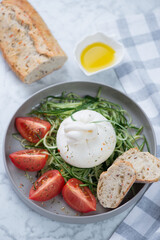 Grey plate with burrata, cucumber and tomato salad, vertical shot on a light-grey marble background