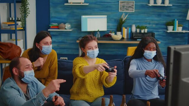 Mixed race women with protection mask playing video game holding wireless controller enjoying new normal party relaxing with friends. Diverse people keeping social distancing against global pandemic