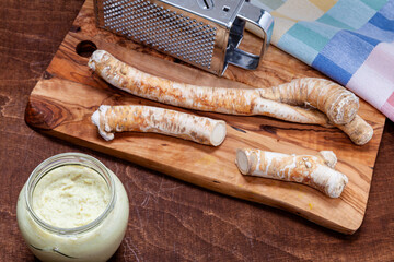 Grated horseradish dip in glass jar, root of horseradish and metal grater on wooden cutting board.