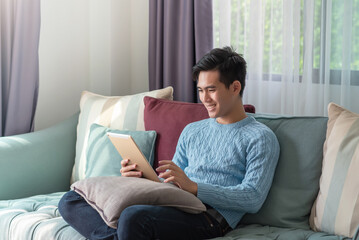 Fototapeta premium Happy young asian man sitting on the sofa watching a tablet at home.