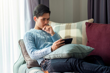 Obraz premium Leisurely day of young Asian man wearing headphones watching their phone sleep at sofa at home.