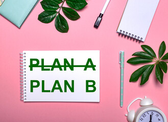 PLAN B is written in green on a white notepad on a pink background surrounded by notepads, pens, white alarm clock and green leaves. Educational concept