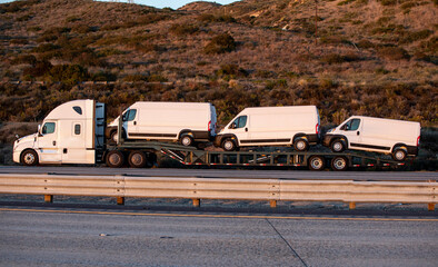 Three new white vans being transported on a flatbed truck in afternoon light. The transport truck...