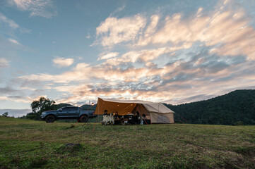 Group of friends relaxing inside large tent and pickup parked on hill in countryside at evening - obrazy, fototapety, plakaty
