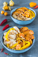 Two Buddha bowls with chicken, rice, bell pepper, and sweet potato, small bowls with minced peanuts and spicy sauce, trend and healthy food on gray background, vertical