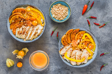 Two Buddha bowls with chicken, rice, bell pepper, and sweet potato, small bowls with minced peanuts and spicy sauce, trend and healthy food, top view on gray background