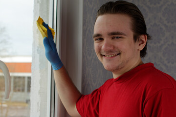 A young guy in a red T-shirt and blue gloves holds a yellow rag in his hands and wipes the window. Window cleaner concept