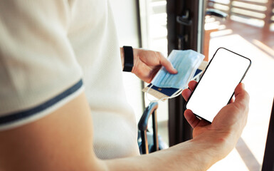 The man holding the mobile phone with empty space as a mockup for digital vaccination certificate in one hand and passport, mask and ticket in another one