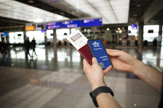 IATA Travel Pass Initiative. Phone in airport and airplane in the background. Passport and flight tickets. Hand holding mobile COVID-19 app for vaccination