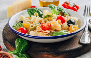 Heart pasta salad with colorful tomatoes, olives, parmesan and basil leaves on gray background. Italian traditional food. Selective focus
