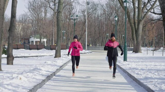 Man and woman wearing pink and black sportswear running in winter park in slow motion. Tracking shot fit couple enjoying outdoor activities together. Concept of sport