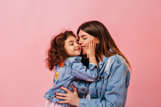 Relaxed preteen girl embracing with mother. Winsome young mom kissing daughter on pink background.