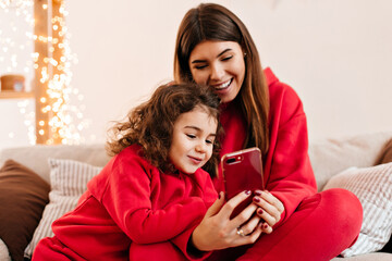Carefree brunette woman with daughter using smartphone. Indoor shot of family in red clothes sitting on couch.