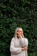 blonde woman in sweater, standing against background of wall of green leaves, hands near her face, eyes closed.