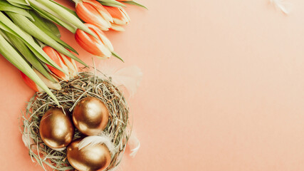 Easter eggs gold. Golden colour egg in basket with spring tulips, white feathers on pastel pink background in Happy Easter decoration. Congratulatory easter design. Flat lay, top view.