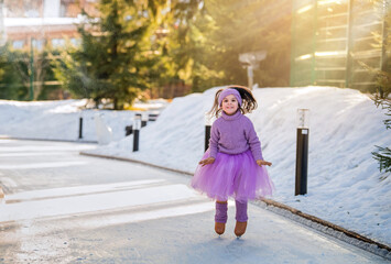 little girl in pink sweater and full skirt rides on sunny winter day on an outdoor ice rink in park