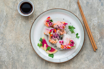 Bright spring rolls with watermelon radish, rice noodles, carrots and shrimps on a clay plate on a gray concrete background. Asian food.