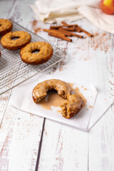 Close Up of Homemade Maple-Glazed Apple-Cinnamon Doughnut on a White Wood Table; Unglazed Doughnuts on a Wire Rack, Apple,  and Cinnamon Sticks in Background