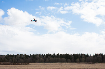 A small plane flies over the field and forest. Sky with clouds. Small airfield in the Leningrad region, St. Petersburg, Russia. The plane leaves a plume of smoke.