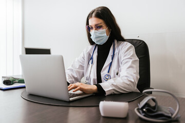 Fototapeta na wymiar Young doctor working on laptop in office analyzes medical records of his patients infected whit Coronavirus Covid-19 wearing protective face mask - Professional sitting at desk looks at the screen