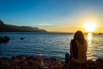 A young and beautiful girl dressed in casual clothes looking at the sunset at a beautiful beach in Artà Mallorca Balearic Islands Spain with a impressive view of the sea and the Tramuntana mountains