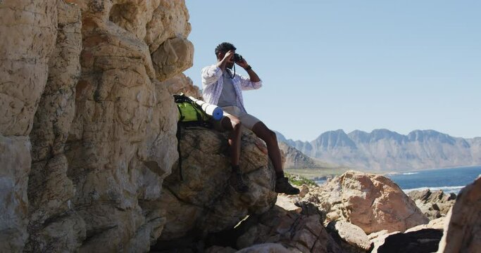 African american man hiking sitting on rock taking photos by the coast