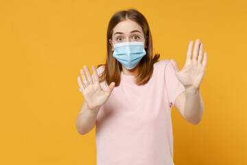 Young woman in basic pastel pink t-shirt, blank print design in sterile face mask, coronavirus covid-19, pandemic quarantine cover with hands show palms isolated on yellow background studio portrait
