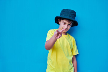Portrait of defiant boy in yellow t-shirt and hat doing, on blue background. Copy space
