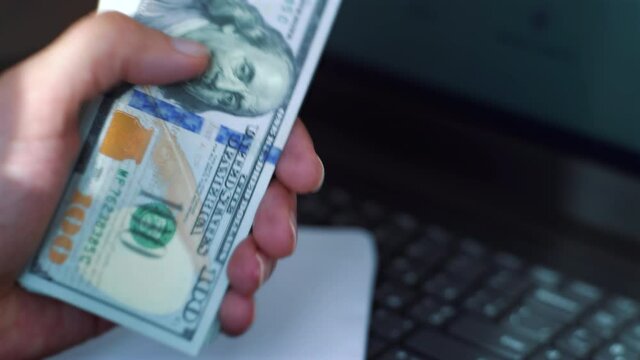 Male hands counting US dollars and putting them in envelope, laptop on background. Closeup businessman getting salary in cash. Concept of money