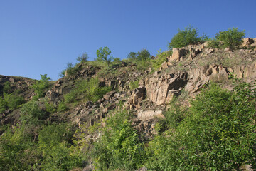 Fototapeta na wymiar A rocky slope of an abandoned granite quarry in the Nikolaev region of Ukraine on a hot summer day. Sharp stones overgrown with sparse vegetation. Blue cloudless sky over the cliffs.