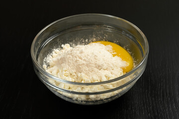 Grainy cottage cheese, egg, flour in a glass bowl on a black table close-up