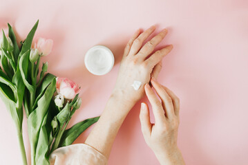 Female hands smearing moisturizer on a pink background with pink fresh spring tulip flowers. Skin care, beauty, flatlay