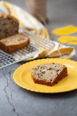 Fototapeta na wymiar A Slice of Banana Nut Bread on a Yellow Plate with Loaves on Cooling Rack in Background; Twine and Banan Nut Bread Tags in Background; Gray Background