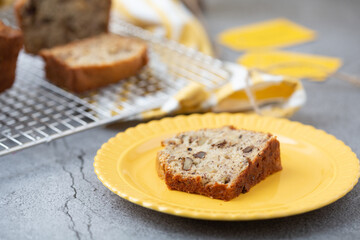 A Slice of Banana Nut Bread on a Yellow Plate with Loaves on Cooling Rack in Background; Twine and Banan Nut Bread Tags in Background; Gray Background