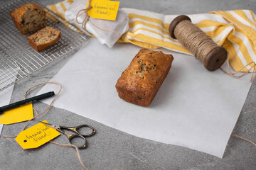 Loaf of Homemade Banana Nut Bread on Parchment with Loaf Sliced on Cooling Rack in Background; One Wrapped in Parchment Paper with Tag in Background; Tags and Twine in Background