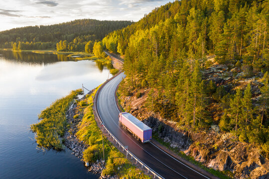 Aerial view of semi truck with cargo trailer on road curve at lake shore with green pine forest. Transportation background. Beautiful nature landscape at sunset in Republic of Karelia, Russia