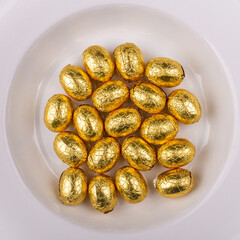 Top view of a pile of yellow golden, wrapped in shiny tinfoil, chocolate shiny Easter eggs in a white round bowl 