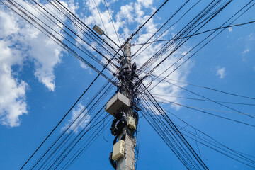 City power line pole with a lantern on top. Numerous wires are connected to the post. Pole is hung with mass of switch boxes and coils of cables