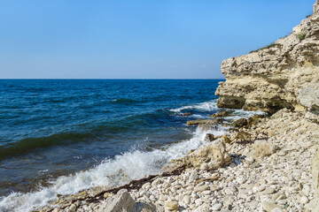 Seascape with cliff broken by endless waves, pebble beach and small silhouette of ship on far horizon