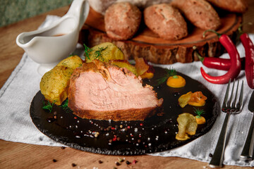 Meat with baked potatoes with dill, sauce and mustard on a black slate dish next to chili peppers and buns and bread on a wooden board.