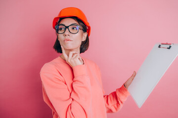 Pretty thoughtful  woman worker builder hold white sign board blank against pink background. Building helmet.