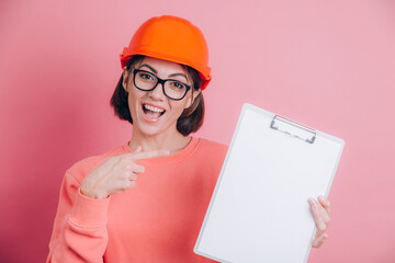 Smiling woman worker builder hold white sign board blank against pink background. Building helmet. Point index finger.